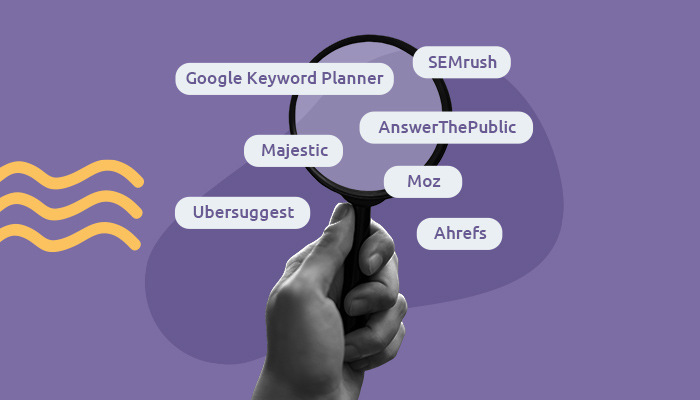 Imagem: uma mão segura uma lupa, sobre a qual circulam o nome de ferramentas de palavras-chave: Google Keyword Planner, SEMrush, Majestic, Ubersuggest, AnswerThePublic, Moz e Ahrefs.